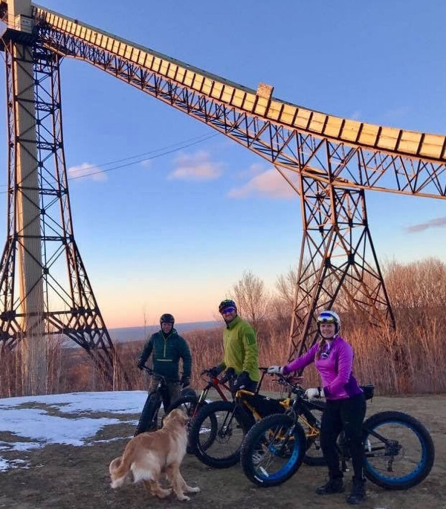 Three bicyclists smile at a vantage point with a view of Copper Peak's ski jump.